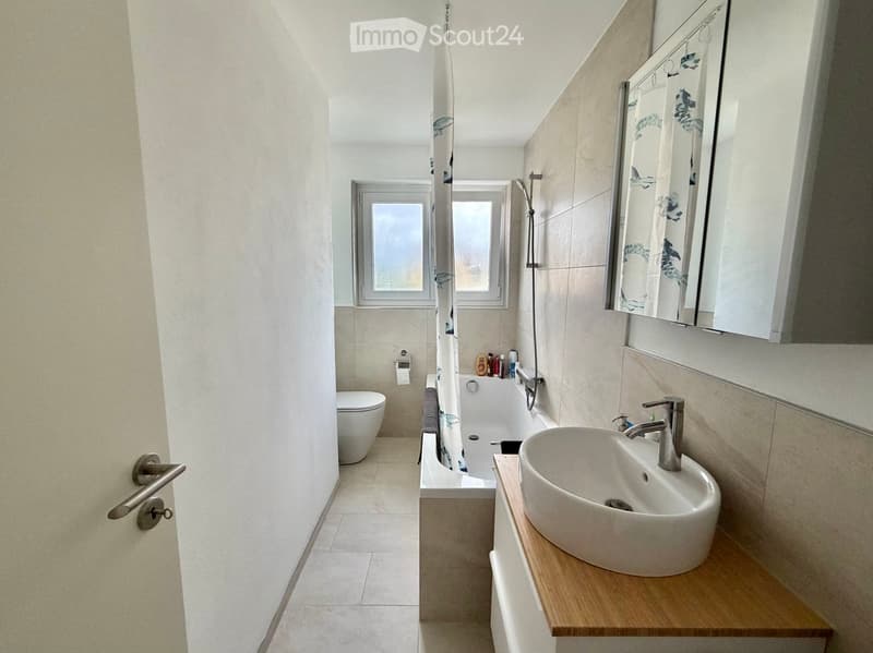 Modern bathroom with a white sink, bathtub, toilet, and a large mirror above the sink. The space features light-colored tiles and a wooden vanity.