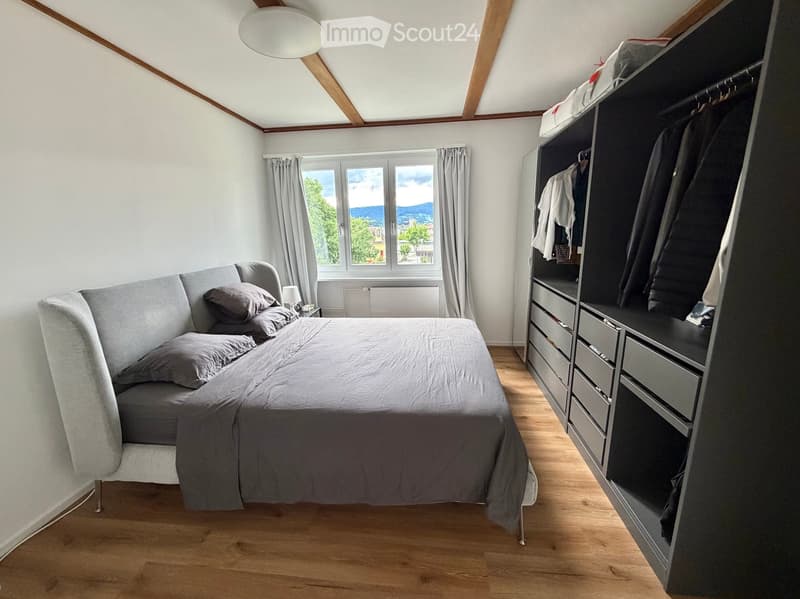 Modern bedroom with a grey upholstered bed, matching pillows, a large built-in wardrobe with drawers, and a window with curtains offering a scenic view.
