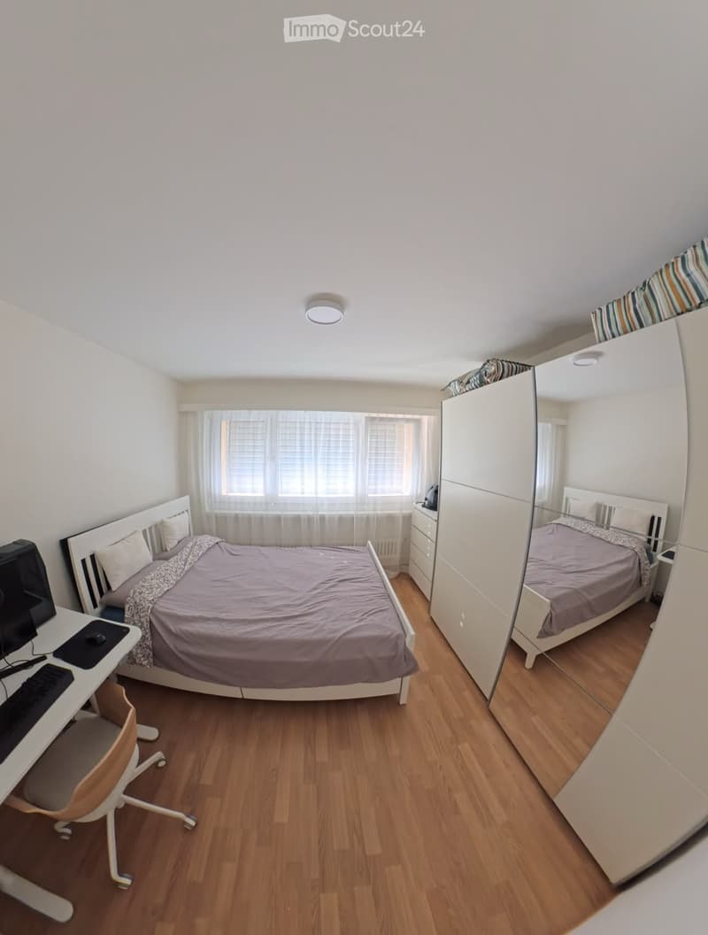 A modern bedroom with a large bed, a desk with a laptop, a chair, a wardrobe with a mirror, and a window with blinds.