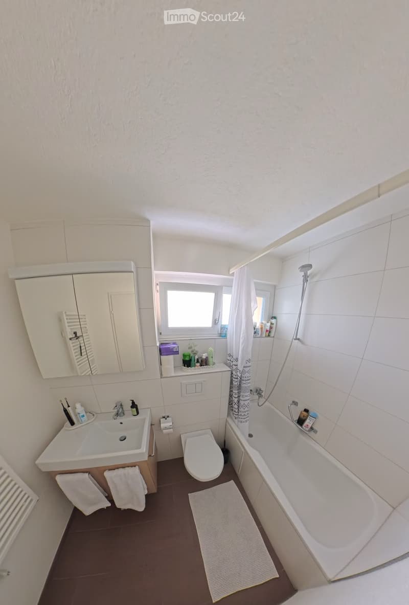 Modern bathroom with white tiles, a sink, toilet, bathtub, mirror, and a window. Includes towels, a shower curtain, and various toiletries on the sink and bathtub ledge.