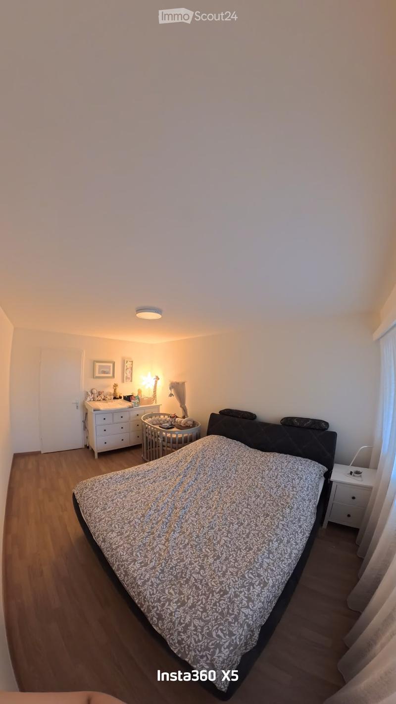 A well-lit bedroom with a large bed covered in a patterned duvet, a nightstand with a lamp, a drawer unit, and a small baby crib in the corner.