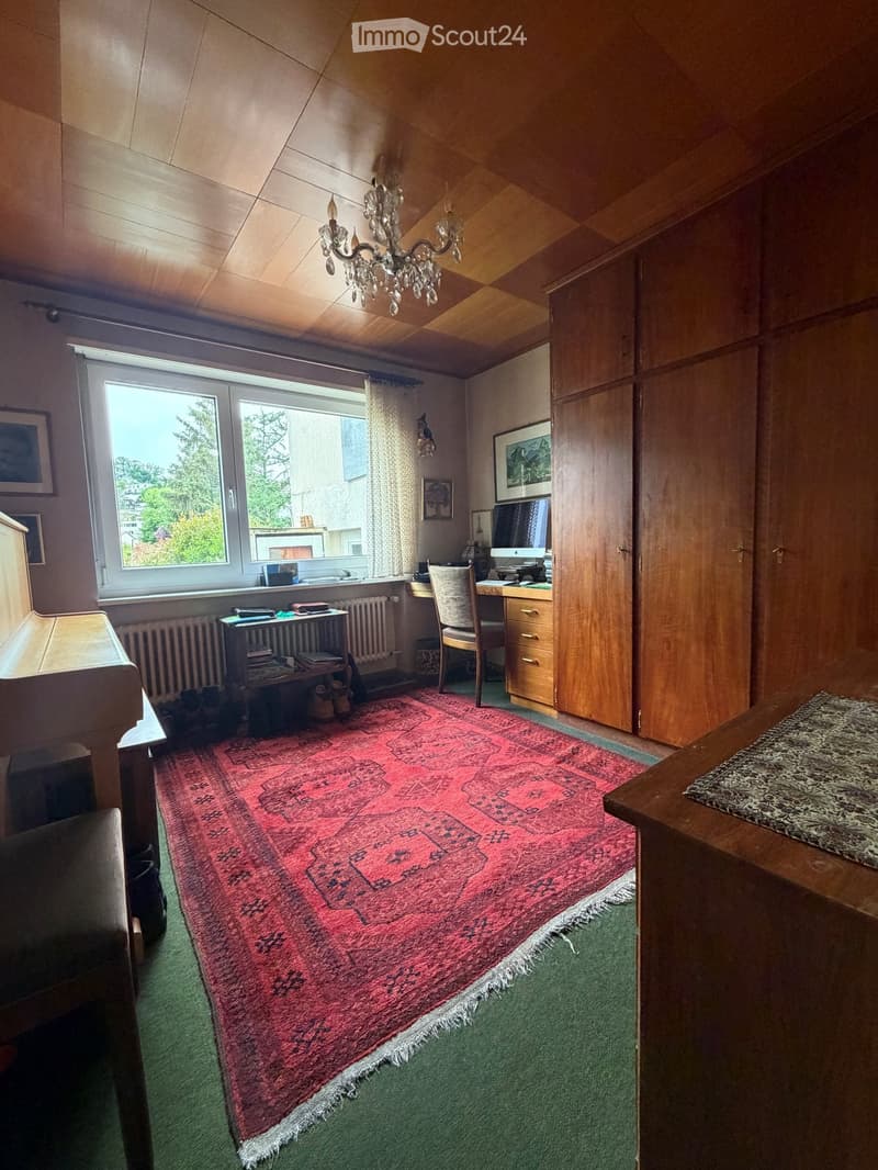 A cozy room with wooden walls and ceiling, featuring a large red patterned rug, a desk with a computer, a chair, a piano, and a chandelier hanging from the ceiling.