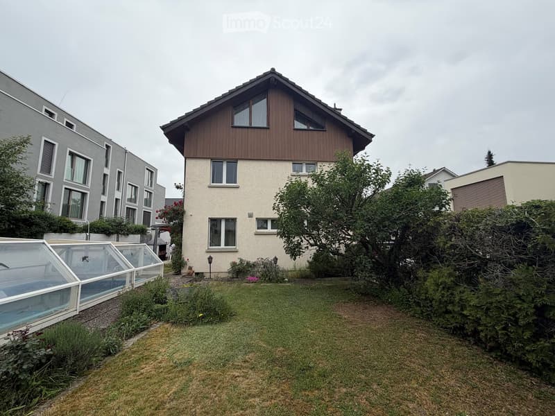 A well-maintained garden with a lawn, a swimming pool covered by a glass enclosure, and surrounding trees and shrubs. The house in the background has a wooden facade and multiple windows.