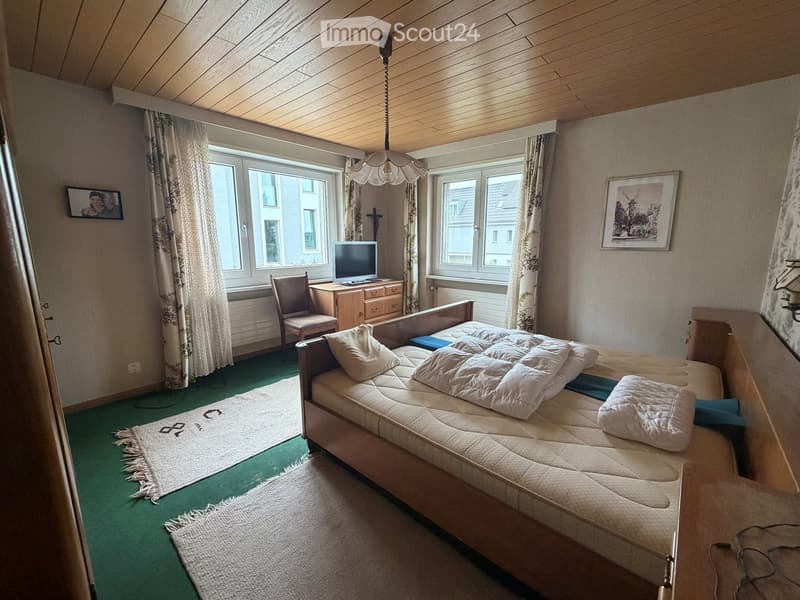 Cozy bedroom with a double bed, nightstand, chair, dresser, and television. Green carpeted floor, wooden ceiling, and large windows with curtains.