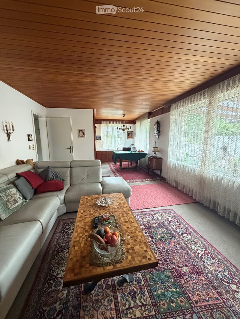 A cozy living room with a sectional sofa, coffee table, dining table, and various decorative items. The room features wooden paneling on the ceiling and walls, large windows with curtains, and several area rugs.