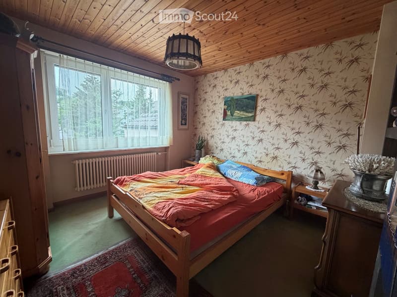 Cozy bedroom with wooden bed, colorful bedding, nightstand, lamp, picture frame, and potted plant on windowsill.