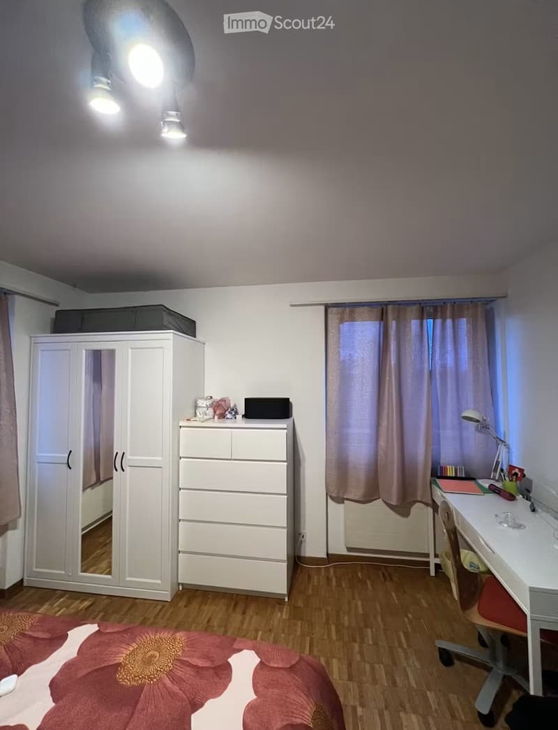 Well-lit bedroom with wooden flooring, a large wardrobe, a drawer unit, a bed with a floral duvet, a desk with a chair, and a lamp on the desk.