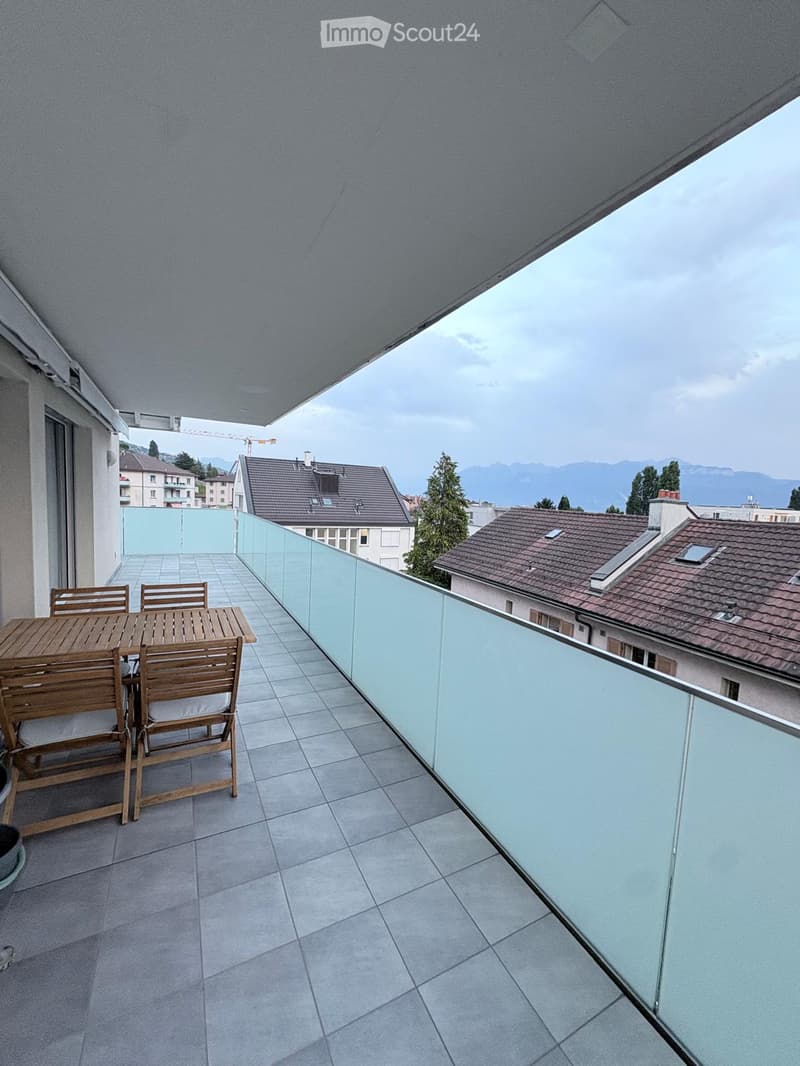 Balcony with tiled floor, glass railing, wooden table and chairs, overlooking cityscape and mountains.