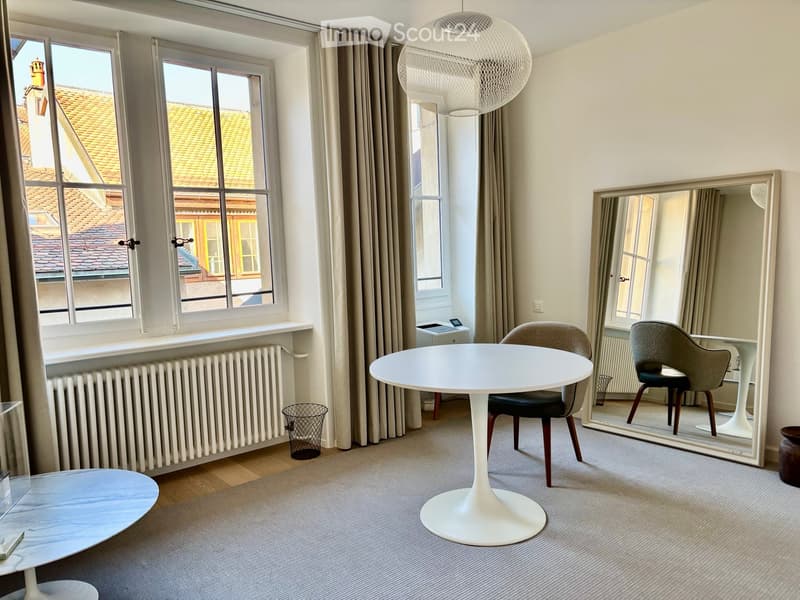 Modern, minimalist room with large windows, a round white table, two chairs, a large mirror, and a hanging spherical lamp.