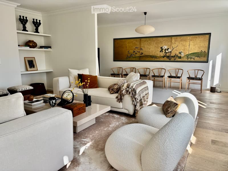 Modern living room with white walls, light wood flooring, and a large abstract painting. Features a white sectional sofa, a round ottoman, a coffee table with books and a vase, and several wooden chairs arranged around a rug.