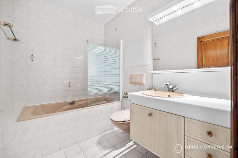 Modern bathroom with white tiles, a bathtub, a sink with a cabinet, and a toilet. Features a glass shower enclosure and a wooden door.