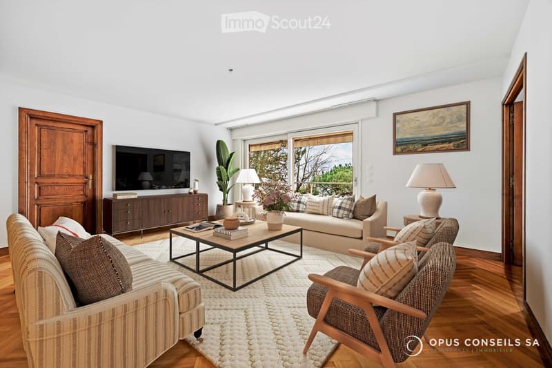 Bright living room with wooden flooring, large windows, and a view. Features a beige sofa, coffee table, armchair, table lamps, and a TV on a wooden cabinet.