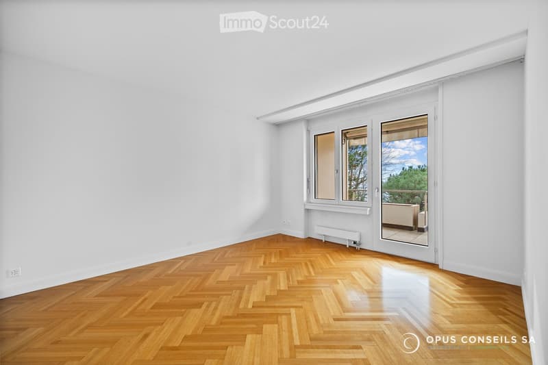Empty room with white walls and herringbone wooden flooring, featuring large windows and a sliding glass door leading to a balcony.