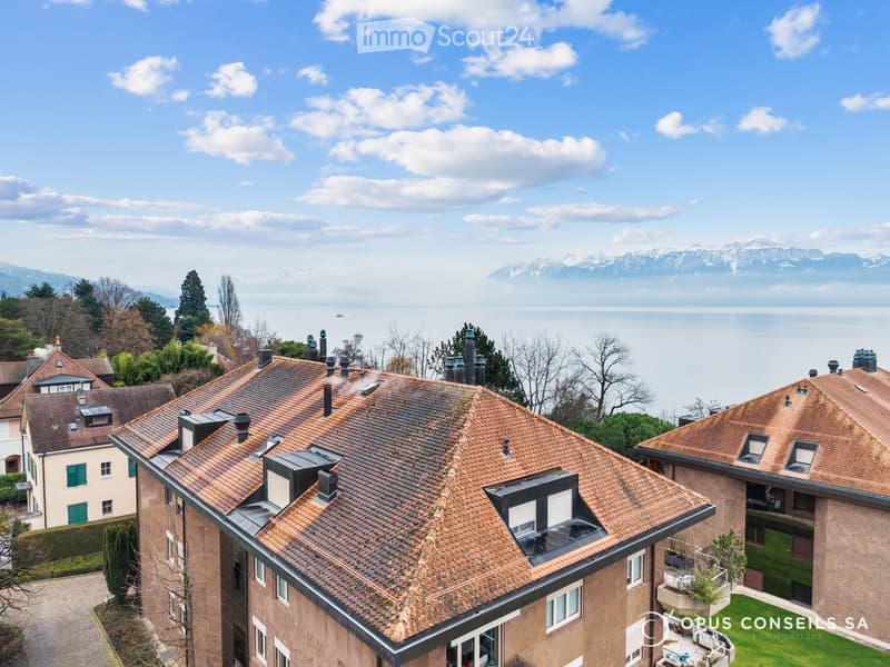 A multi-story residential building with red-tiled roofs, surrounded by trees and overlooking a lake and distant mountains.