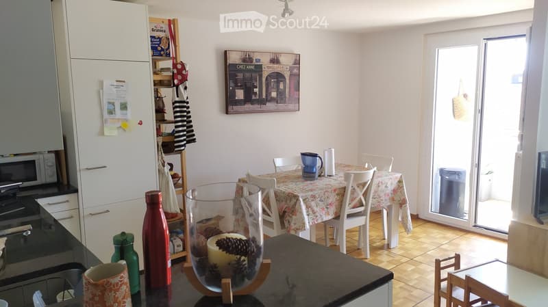 Bright kitchen and dining area with white cabinets, a microwave, oven, and a dining table with chairs. A large window opens to a balcony, and decorative items like a candle and picture frame are visible.