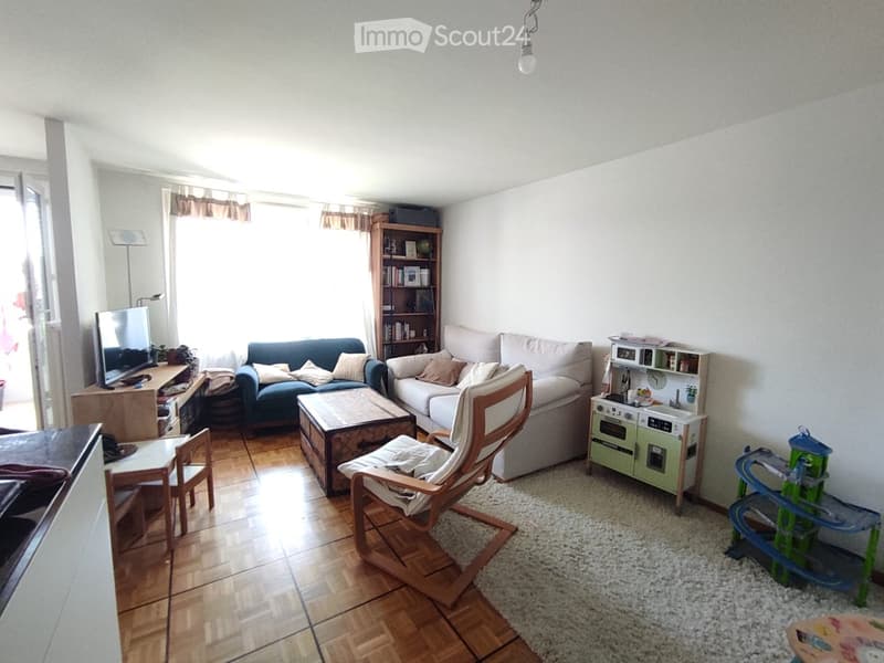 A well-lit living room with wooden flooring, a grey sofa, a coffee table, a wooden chair, a bookshelf, a television, a small toy kitchen, and a toy stroller.
