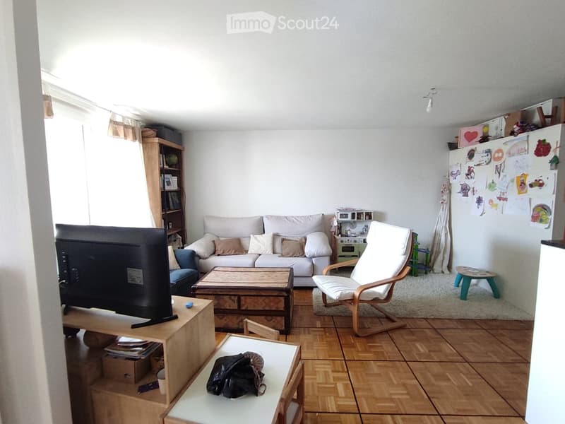 Living room with a sofa, coffee table, chair, television, bookcase, and a small stool. The room features wooden flooring and a white wall with children's drawings.