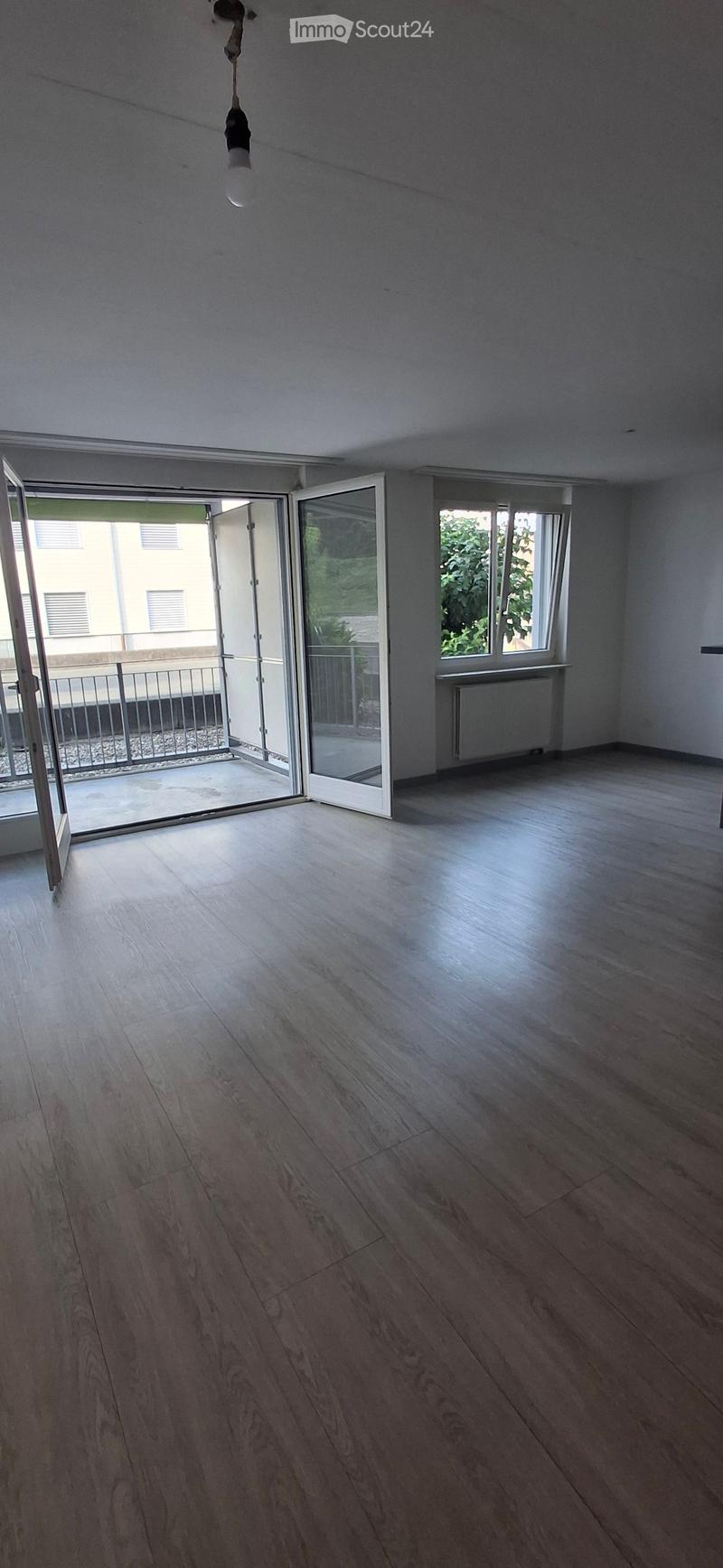 Empty room with wooden flooring, white walls, and large windows. Features a sliding glass door leading to a balcony area.