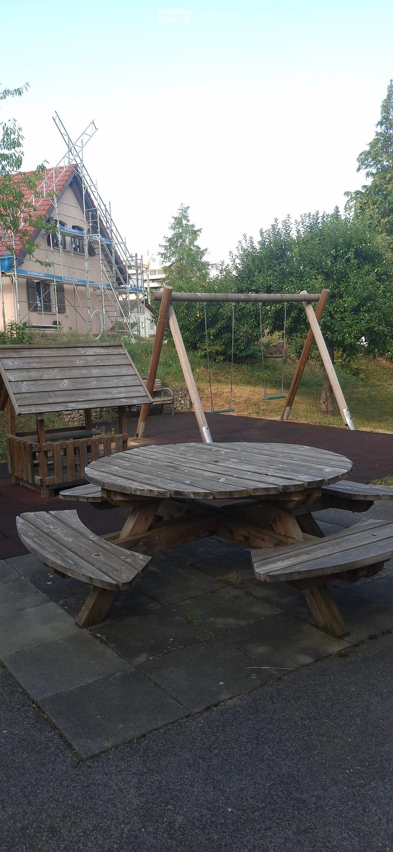 Outdoor area with a wooden picnic table, benches, and a swing set in the background.