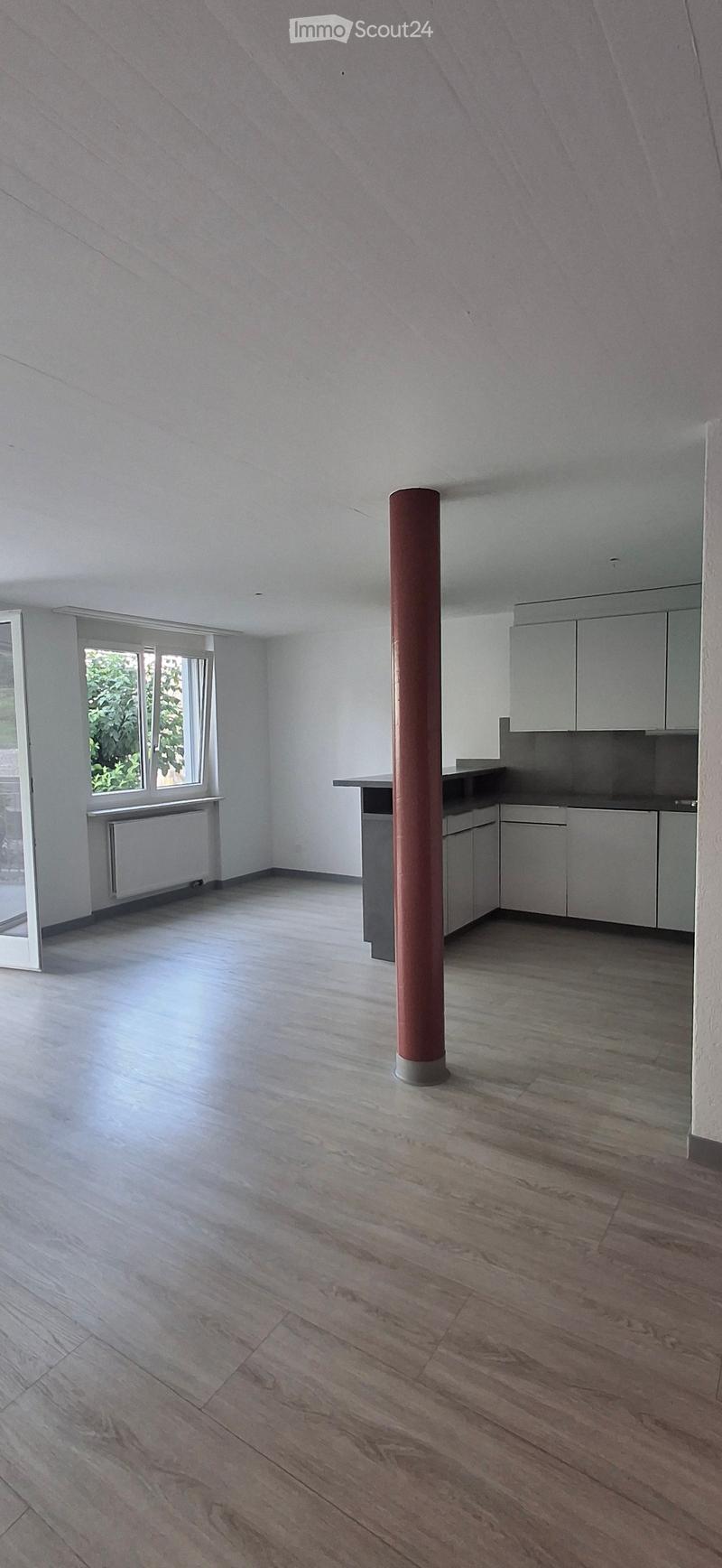 Empty room with wooden flooring, white walls, and a central support column. Features a window with a view of greenery and a small kitchen area with cabinets and a countertop.