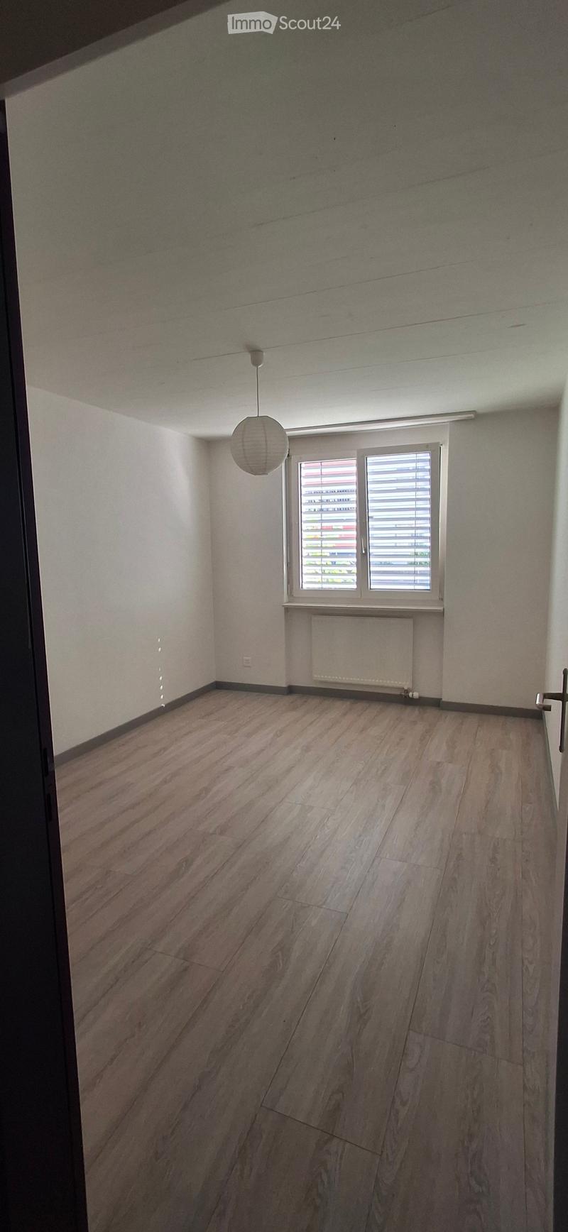 Empty room with wooden flooring, white walls, a single window with blinds, a radiator, and a hanging light fixture.