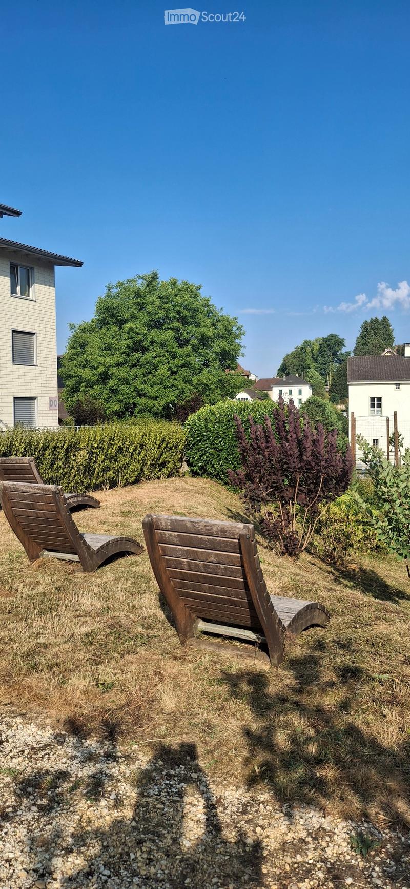 Garden area with two wooden lounge chairs on a gravel and grass surface, surrounded by bushes and trees, with a house visible in the background.
