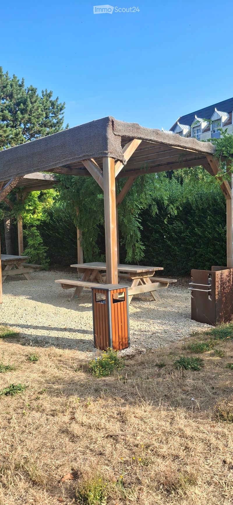 Outdoor seating area with wooden picnic tables, benches, a covered shelter, trash bins, and surrounding greenery.