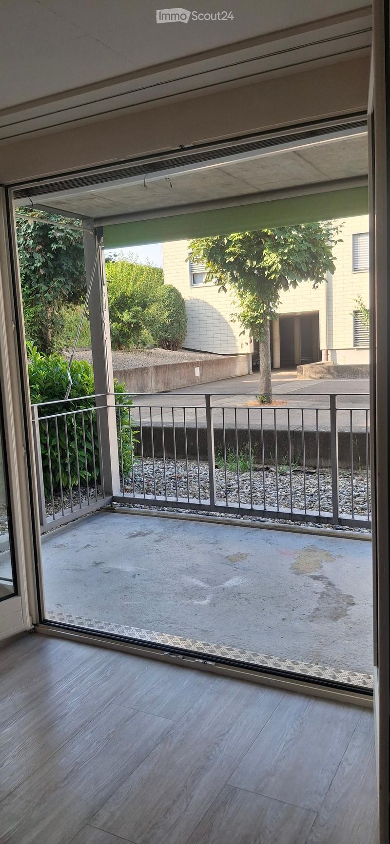 Open balcony with wooden flooring, metal railing, and a view of a garden area with trees and a building in the background.
