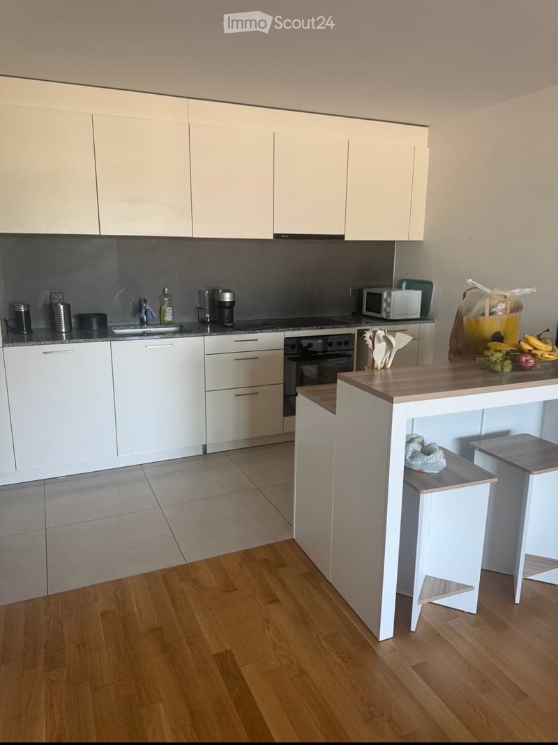Modern kitchen with white cabinets, wooden floor, island with stools, and various appliances including a microwave, coffee maker, and blender. Countertop has a bowl of fruit and a cutting board.