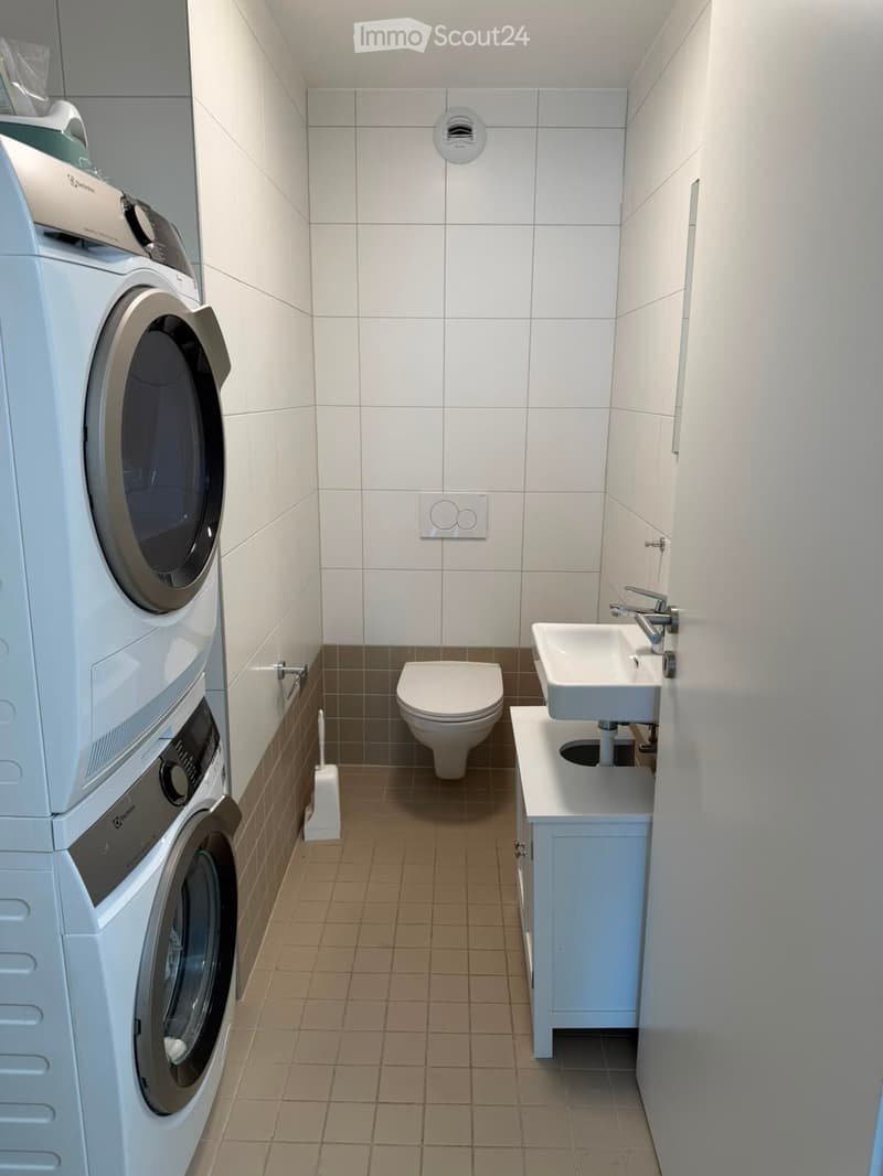 Modern bathroom with white tiles, a toilet, a sink with a faucet, a washing machine, and a dryer. The floor is tiled in a warm brown color, and there is a small cabinet next to the sink.