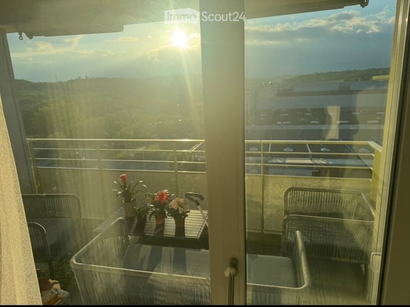 Balcony with a view of the city and surrounding landscape, featuring a table, chairs, and flower pots.