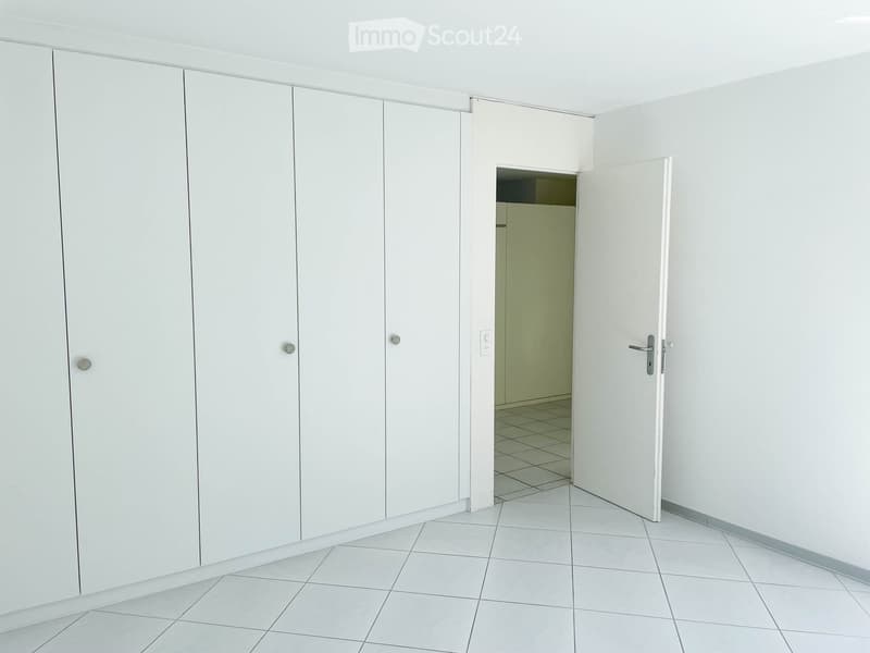 Empty room with white tiled flooring, white walls, and a large open closet with multiple doors. A doorway leads to another room with a tiled floor.