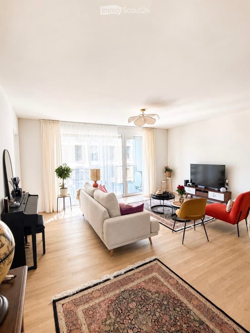 Modern living room with light wooden flooring, large windows with white curtains, a white sofa with colorful cushions, a coffee table, a red armchair, a yellow chair, a piano, a television, and decorative plants.