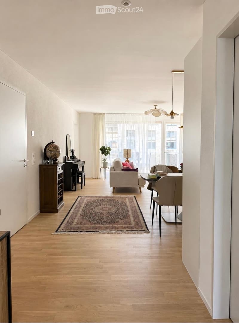 Open-plan living and dining area with light wooden flooring, white walls, large windows with sheer curtains, a patterned rug, a cream sofa with pink cushions, a dining table with chairs, a small piano, a cabinet, and a globe on a side table.