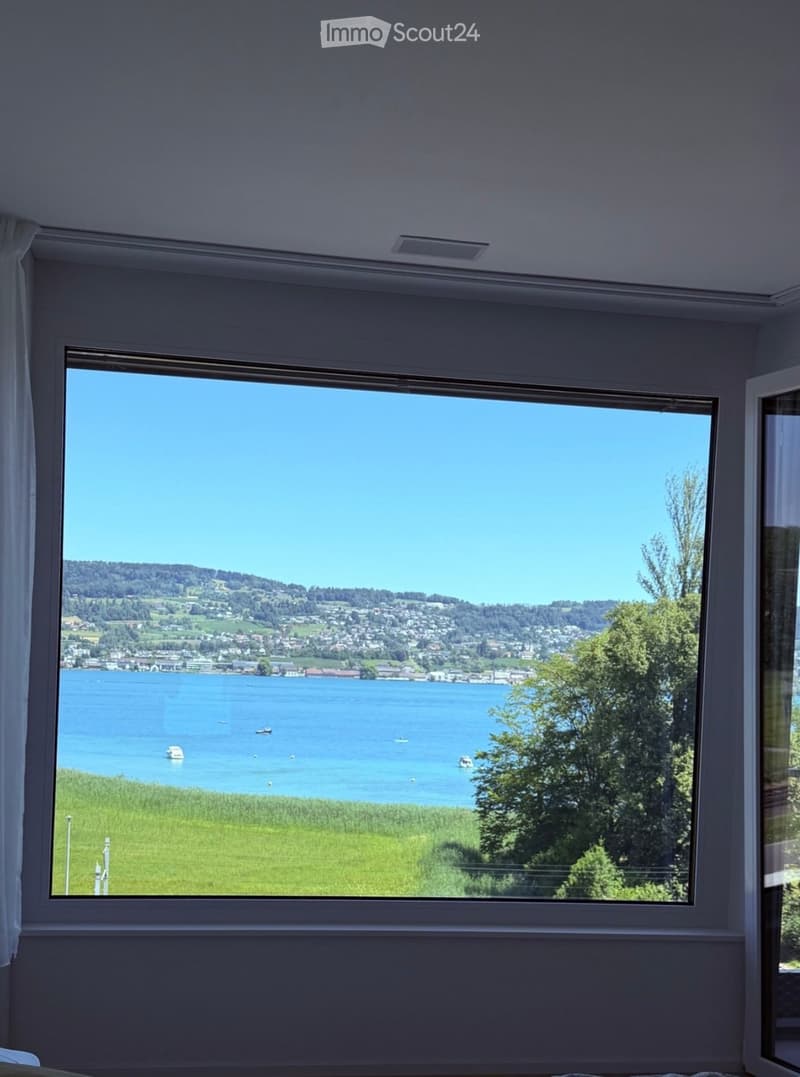 Empty room with large window offering a scenic view of a lake, surrounding greenery, and distant town.