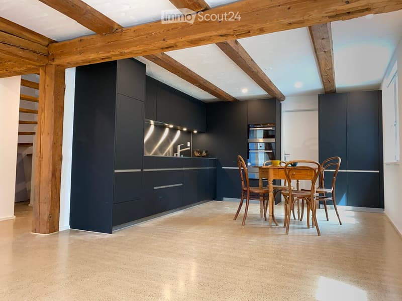 Modern kitchen with dark cabinetry, stainless steel appliances, and exposed wooden beams. Includes a small dining area with a wooden table and chairs.