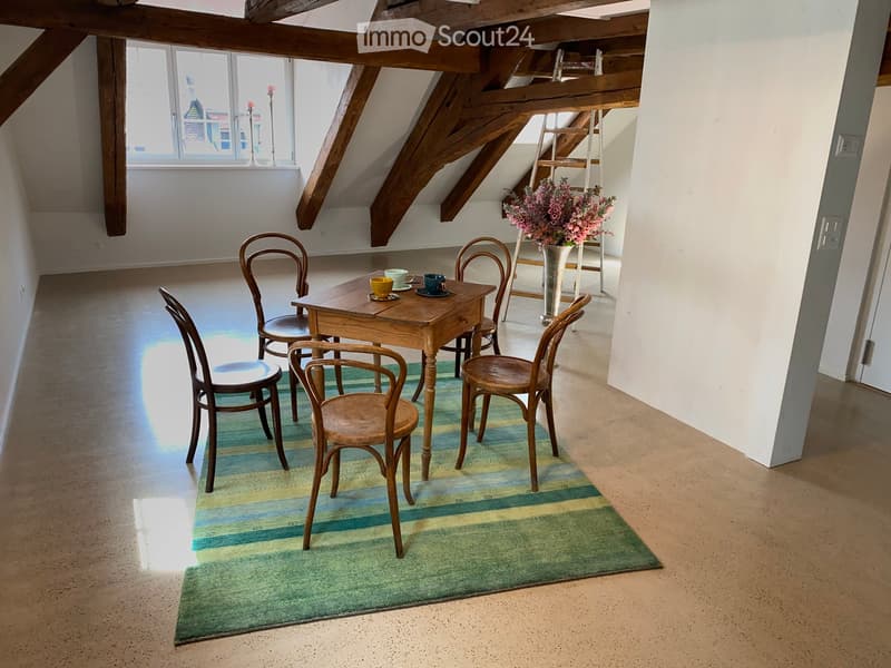 Bright attic room with wooden beams, a small wooden table surrounded by chairs, a vase with flowers, and a colorful striped rug.