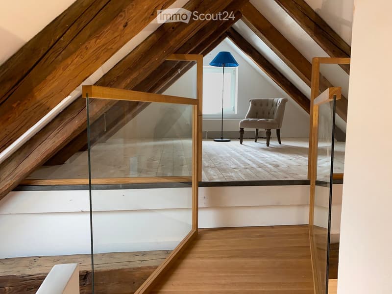 Attic room with wooden beams, glass railing, wooden floor, a single armchair, and a floor lamp near a window.