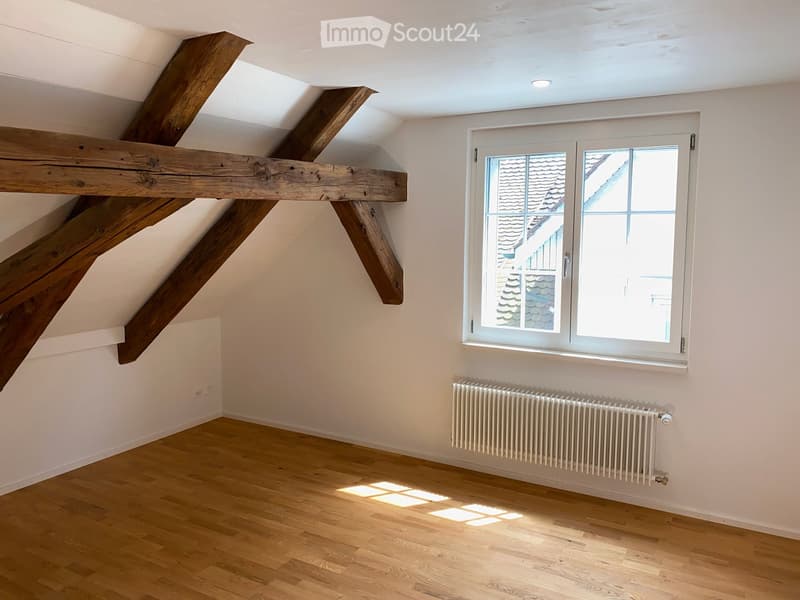 Empty attic room with wooden beams, wooden flooring, white walls, a window, and a radiator.
