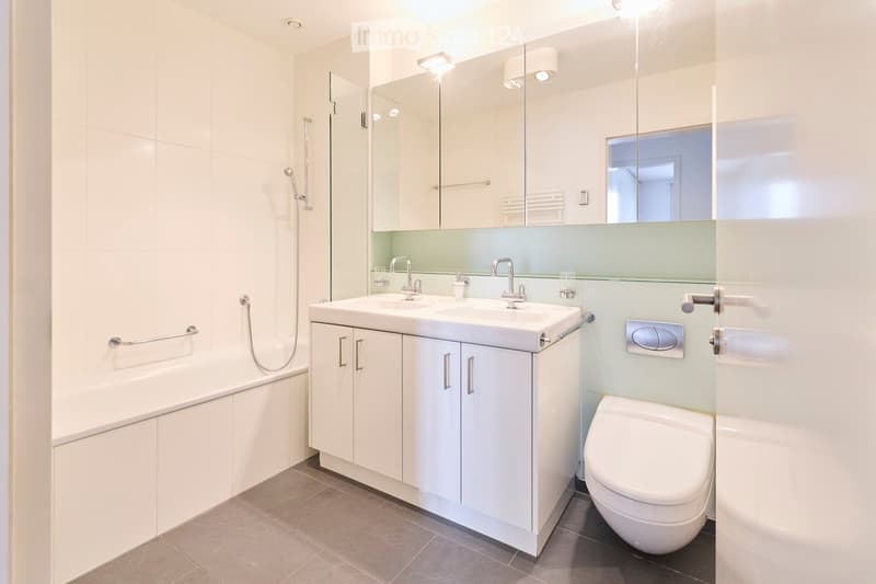 Modern bathroom with white tiles, double sink, toilet, bathtub, and large mirror with built-in lighting.