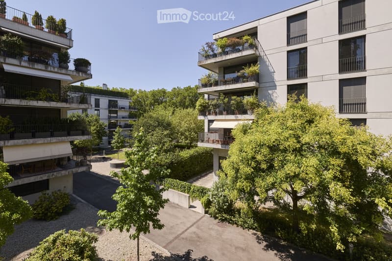 Modern multi-story residential building with balconies, greenery, and trees in the courtyard.