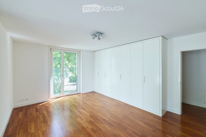 Empty room with wooden flooring, white walls, large sliding glass door leading to a balcony, built-in white closet, and recessed ceiling lighting.