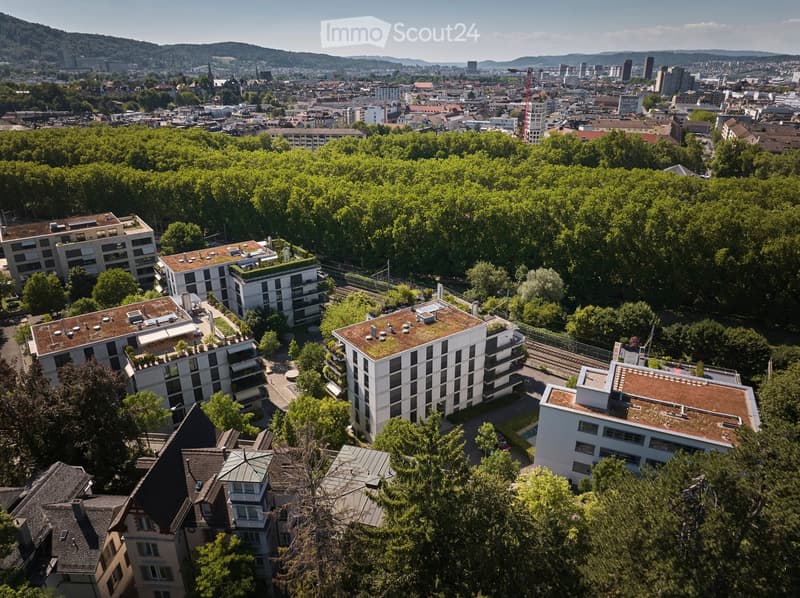 Aerial view of a cityscape with modern apartment buildings, green rooftops, and surrounding trees, with a distant city skyline visible.