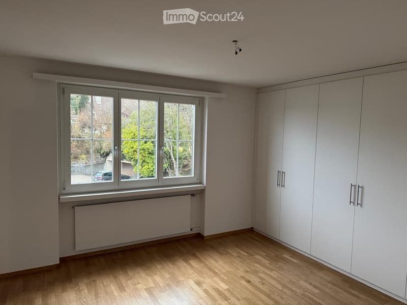 Empty room with wooden flooring, white built-in wardrobes, large window with view of the garden, and radiator below the window.