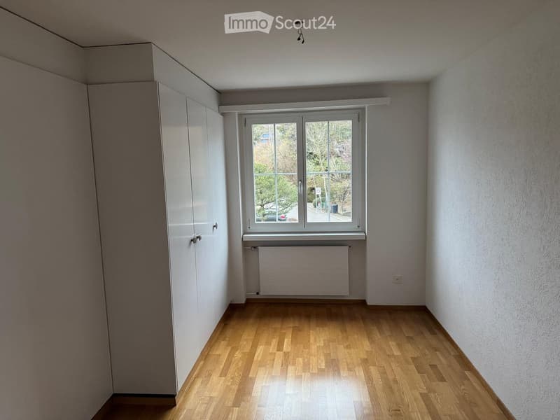 Empty room with wooden flooring, white walls, built-in closet, window with radiator below, and visible outdoor view.
