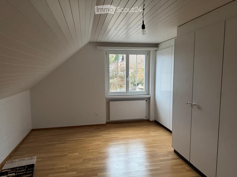 Empty attic room with wooden floors, slanted ceilings, white walls, and a window with a radiator below it.