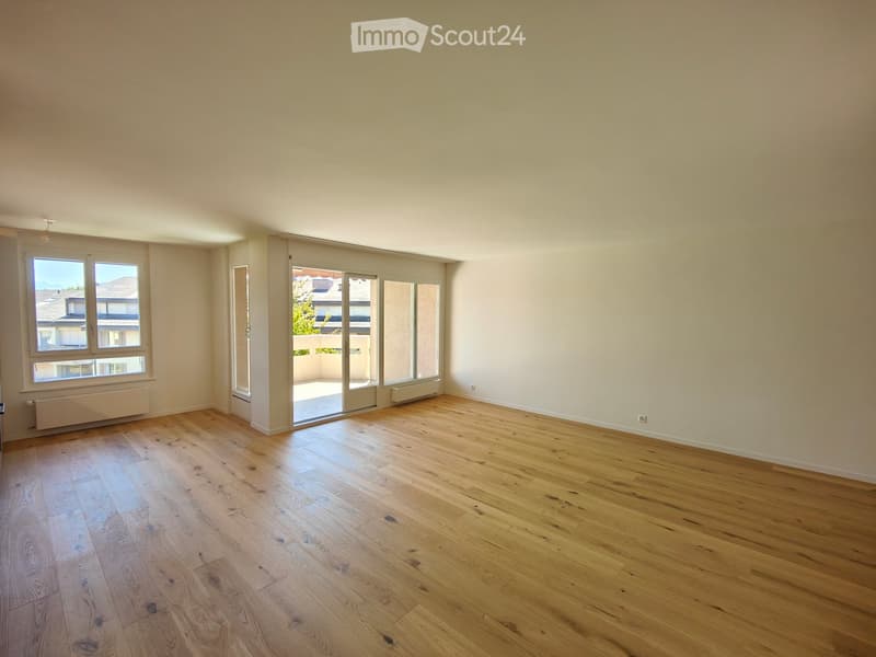 Empty room with light wooden flooring, white walls, large windows, and sliding glass doors leading to a balcony.