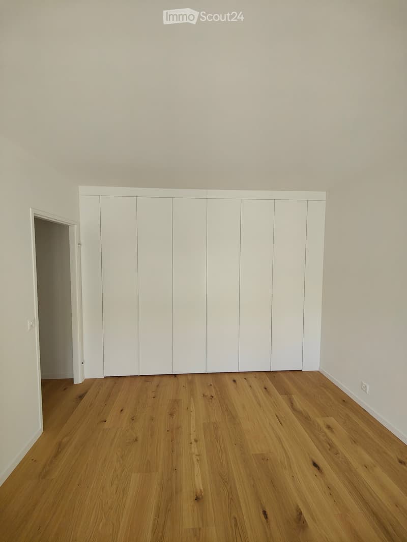 Empty room with light wooden flooring and white walls, featuring a large set of white built-in wardrobes and an open doorway leading to another space.