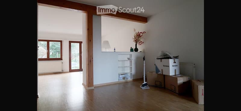 Empty room with wooden flooring, white walls, large windows, and a small built-in shelf unit. Several cardboard boxes are stacked in the corner, and a broom is leaning against the wall.