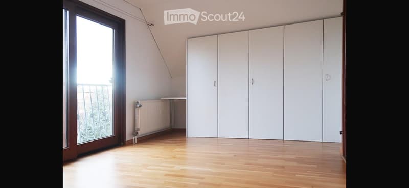 Empty room with wooden flooring, white built-in wardrobes, large window with balcony access, radiator, and a small table.